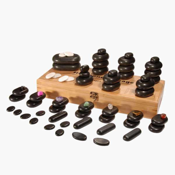A complete 64-piece hot stone massage set arranged on and around a closed wooden storage box, showcasing various stone shapes, sizes, white marble stones, and colorful chakra gems.