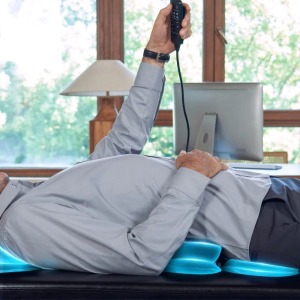 A side view of a client lying on a black massage table, holding a remote control while the table's blue illuminated airbags provide active stretching along their back.